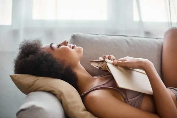 Seitenansicht einer jungen afrikanisch-amerikanischen Unterwäsche, die mit aufgeschlagenem Buch auf einem bequemen Sofa liegt — Stockfoto
