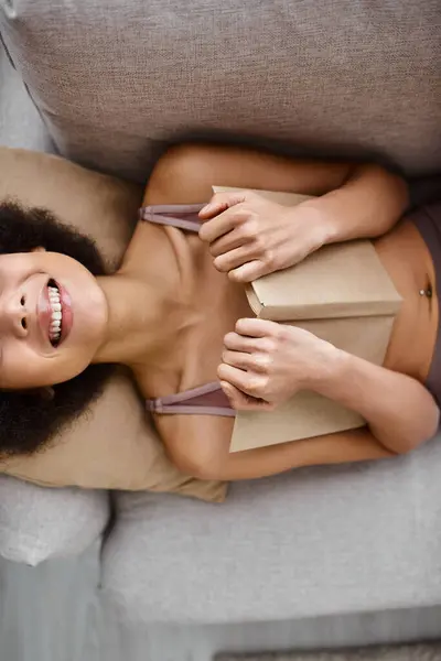 Vista superior da mulher africana feliz cortada roupa interior deitado com livro aberto em um sofá confortável — Fotografia de Stock
