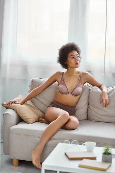 Verträumte junge afrikanisch-amerikanische Frau mit lockigem Haar lümmelt in Dessous auf einem Sofa zu Hause — Stockfoto