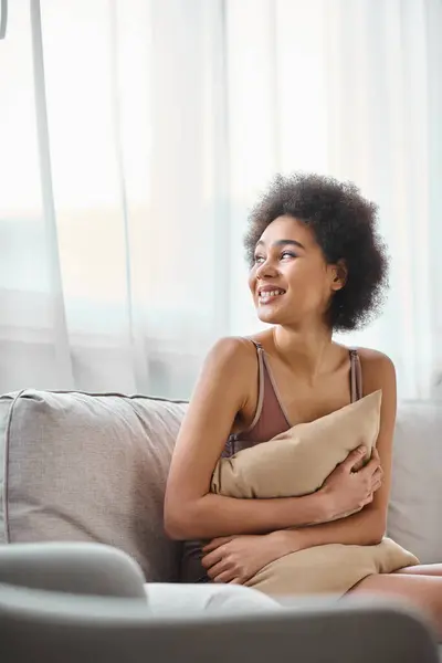 Giovane donna afroamericana dai capelli ricci che si rilassa sul divano in lingerie, sorridendo e distogliendo lo sguardo — Foto stock