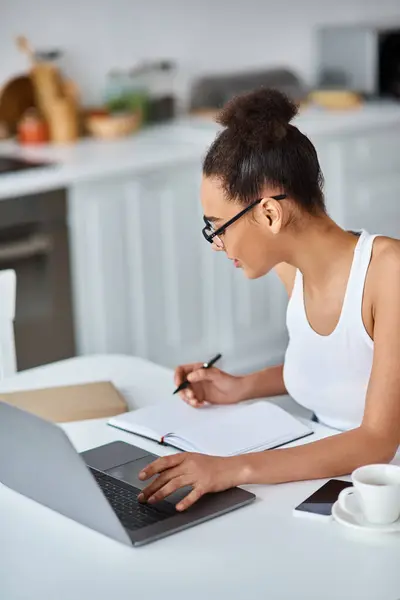 Молодая африканская американка в очках, работающая из дома дистанционно на ноутбуке, записывающая — стоковое фото