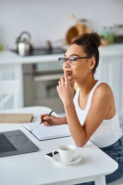Allegra donna afroamericana in occhiali che lavora da casa a distanza sul suo computer portatile, prendendo appunti — Foto stock