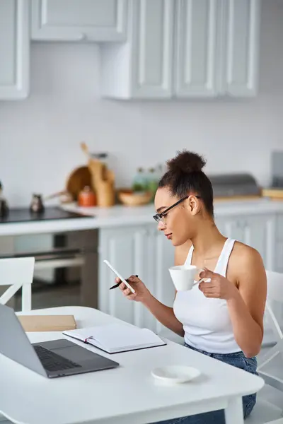 Femme afro-américaine bouclée dans des lunettes de travail de la maison à distance, tenant tasse et smartphone — Photo de stock