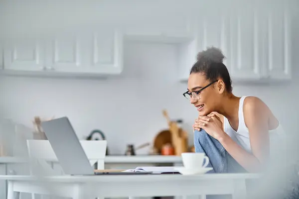 Fröhliche und junge afrikanisch-amerikanische Frau mit Brille, die von zu Hause aus an ihrem Laptop arbeitet — Stockfoto