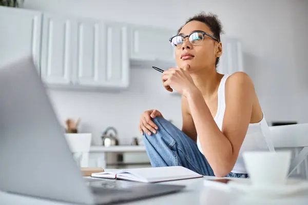 Femme afro-américaine coûteuse dans des lunettes de travail à distance de la maison sur son ordinateur portable, tasse de café — Photo de stock