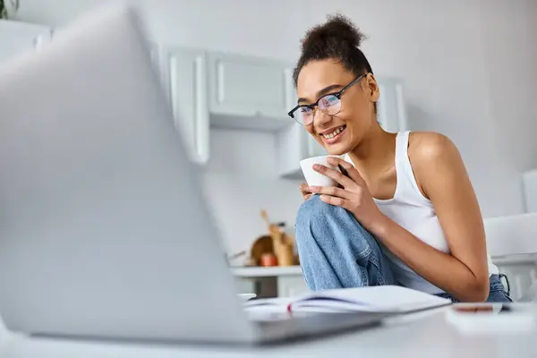 Щаслива афроамериканка в окулярах тримає чашку і працює з дому, використовуючи ноутбук на столі — стокове фото