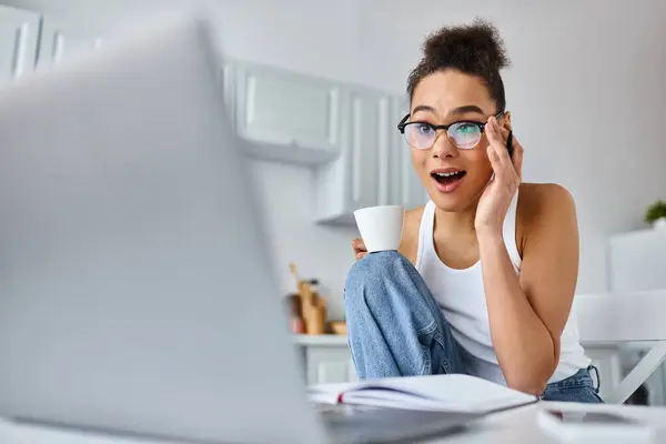 Donna afro-americana stupita in bicchieri in possesso di tazza e guardando il computer portatile sulla scrivania, lavoro a distanza — Foto stock
