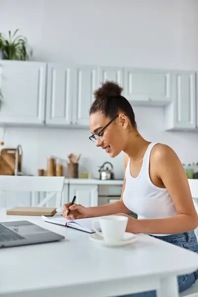 Heureuse femme afro-américaine dans des lunettes prenant des notes près d'un ordinateur portable et une tasse de café, travail à distance — Photo de stock