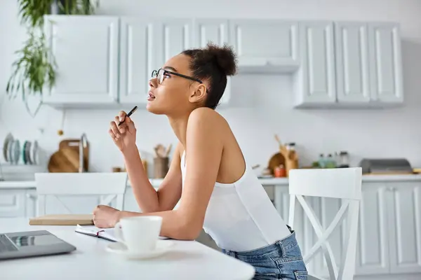 Pensive african american woman in glasses taking notes near laptop and cup of coffee, remote work — Stock Photo