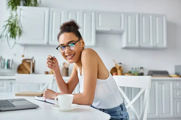 Femme afro-américaine gaie dans des lunettes prenant des notes près d'un ordinateur portable et une tasse de café, travail à distance — Photo de stock