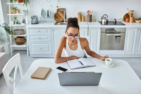 Молодая африканская американка в очках смотрит на свой ноутбук возле чашки кофе, удаленная работа — стоковое фото
