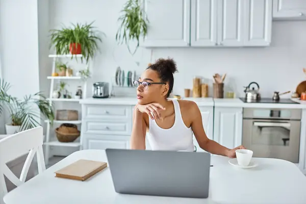 Pensive african american woman in glasses sitting at desk with laptop and cup of coffee, remote work — Stock Photo