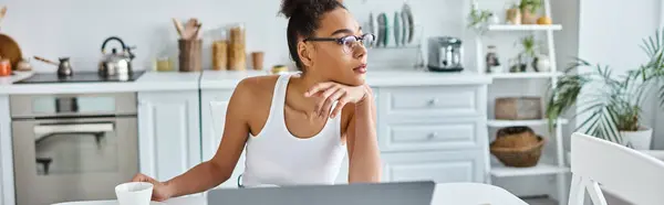 Задумчивая африканская американка в очках сидит за столом с ноутбуком и чашкой кофе, баннер — стоковое фото