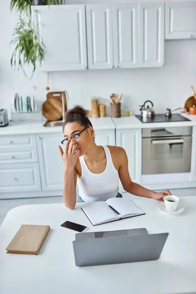 Задумчивая африканская американка регулирует очки и сидит за столом с ноутбуком и чашкой кофе — стоковое фото