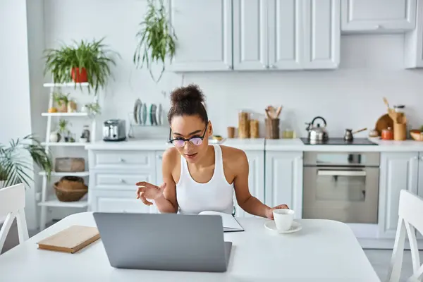 Афроамериканська жінка в окулярах сидить за столом з ноутбуком під час відеодзвінка, віддалена робота — стокове фото