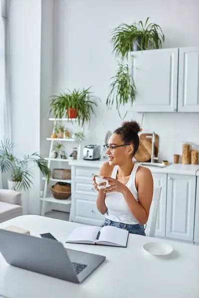 Mulher americana africana alegre em óculos segurando xícara de café perto de laptop na mesa, trabalho remoto — Fotografia de Stock