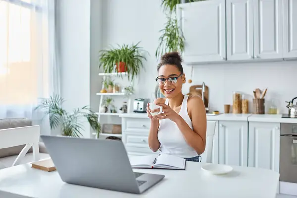 Donna afro-americana positiva in bicchieri che tengono una tazza di caffè vicino al computer portatile sulla scrivania, lavoro a distanza — Foto stock
