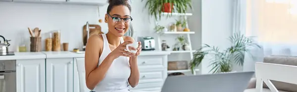 Позитивна афроамериканка в окулярах тримає чашку кави біля ноутбука поруч з нею, банер — стокове фото