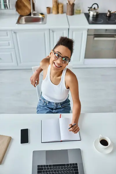 Top view of happy african american woman with pen in hand using laptop next to cup of coffee on desk — Stock Photo