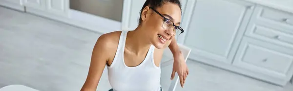 Vista superior de la mujer afroamericana feliz en gafas y camiseta sonriendo en casa, bandera horizontal - foto de stock