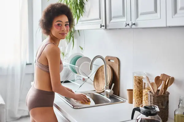Allegra donna afroamericana in lingerie con toppe oculari piatto di lavaggio con spugna in cucina — Foto stock