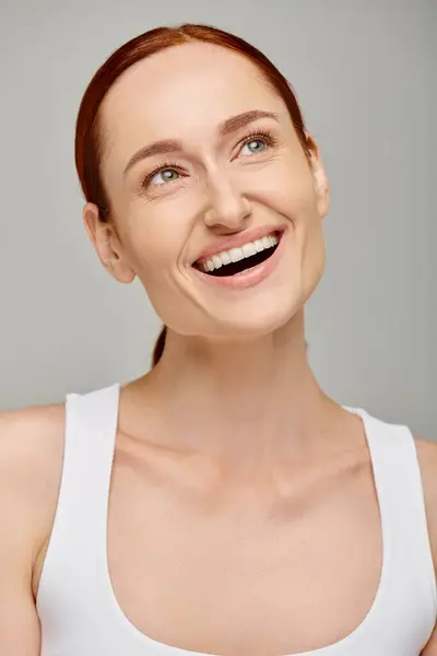 Happy redhead woman in tank top exuding joyful and healthy smile on grey background, looking away — Stock Photo