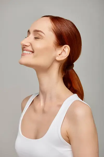 Cheerful redhead woman with closed eyes exuding joyful and healthy smile on grey background — Stock Photo