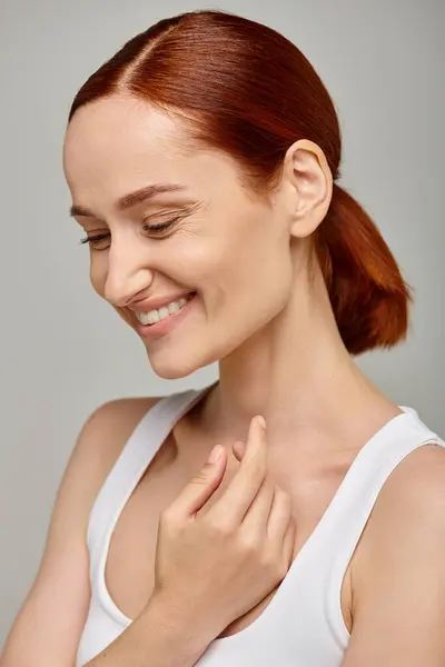 Cheerful redhead woman in white tank top exuding joyful and healthy smile on grey background — Stock Photo