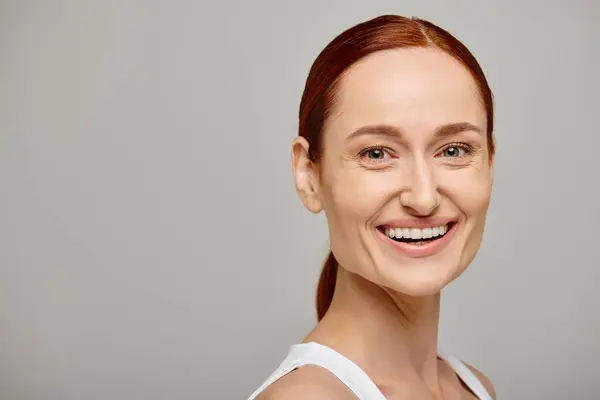 Content and redhead woman in white tank top with a tender healthy smile on a grey background — Stock Photo