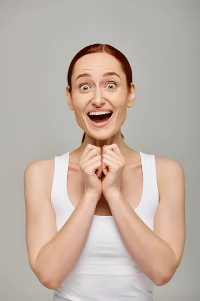 Amazed and redhead woman in white tank top with healthy smile on a grey background, laughter — Stock Photo