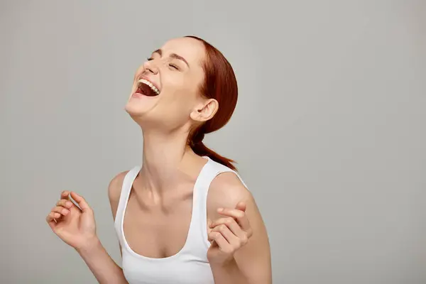 Femme gaie et rousse en débardeur blanc riant un fond gris, sourire radieux et sain — Photo de stock