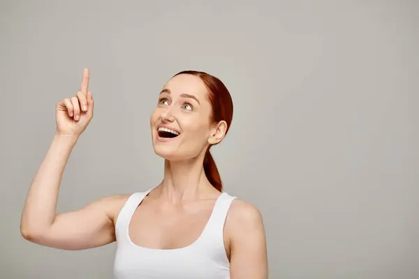 Cheerful and redhead woman in white tank top pointing with finger and looking up on grey backdrop — Stock Photo