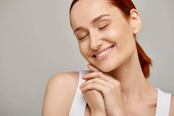 Pleased and redhead woman in white tank top smiling with closed eyes on grey background, tender — Stock Photo