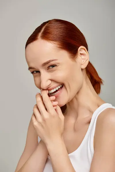 Happy and redhead woman in white tank top laughing and looking at camera on grey background — Stock Photo