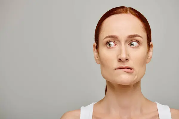 Redhead and worried woman in white tank top embodying concern on grey background, biting lip — Stock Photo