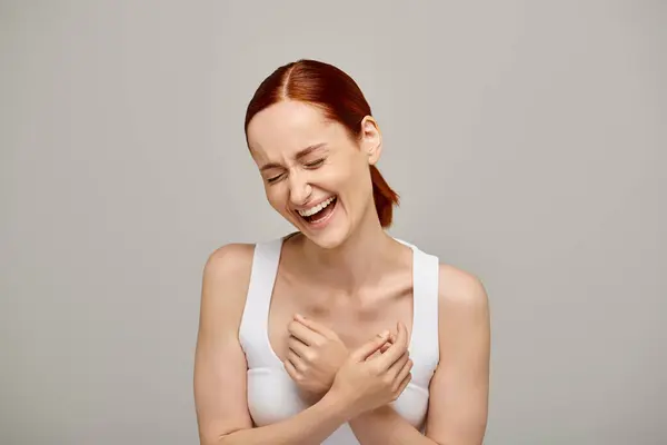 Happy and redhead woman in white tank top laughing with closed eyes on grey background, genuine — Stock Photo