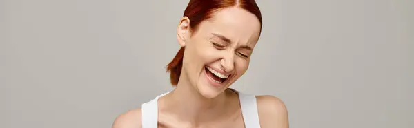 Mujer feliz y pelirroja en camiseta blanca riéndose con los ojos cerrados sobre fondo gris, pancarta - foto de stock