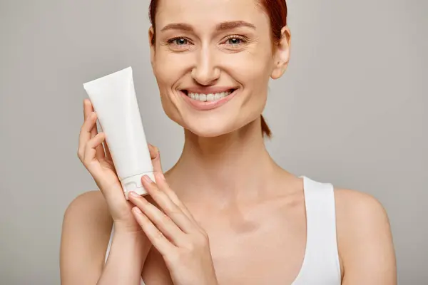 Redhead young and happy woman in 30s holding tube with body lotion on grey background, skincare — Stock Photo