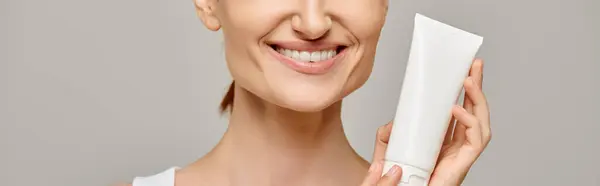 Jeune femme avec un sourire sain tenant tube avec lotion pour le corps sur fond gris, bannière horizontale — Photo de stock