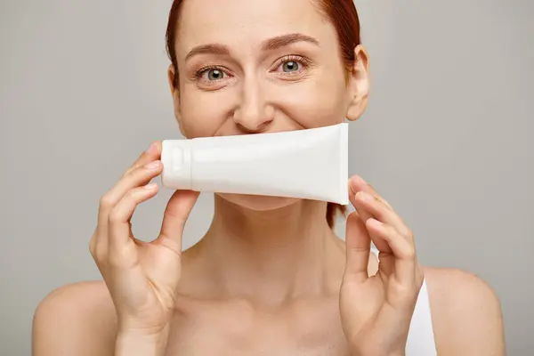 Ruiva jovem de 30 anos cobrindo seu rosto com loção em tubo em fundo cinza, cuidados com a pele — Fotografia de Stock