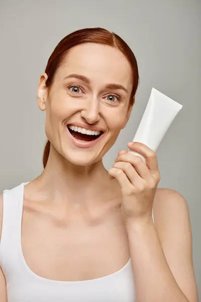 Redhead young and excited woman in 30s holding tube with body lotion on grey background, skincare — Stock Photo