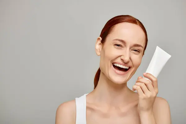 Redhead young and cheerful woman in 30s holding tube with body lotion on grey background, skincare — Stock Photo