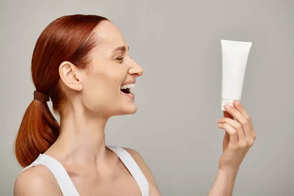 Redhead young and happy woman in 30s looking at tube with body lotion on grey background, skincare — Stock Photo