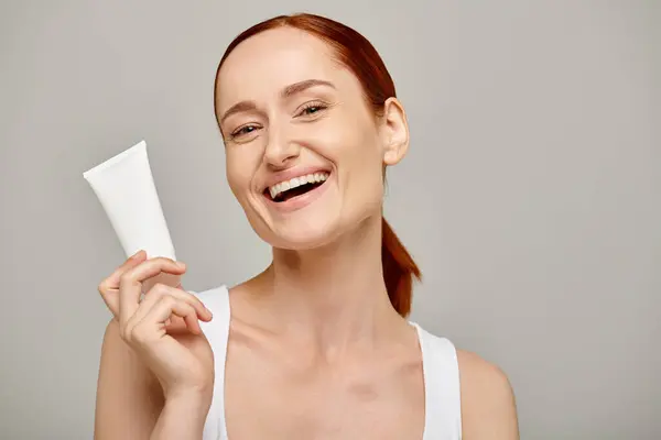 Cheerful young woman with red hair holding tube with lotion on grey background, body care — Stock Photo