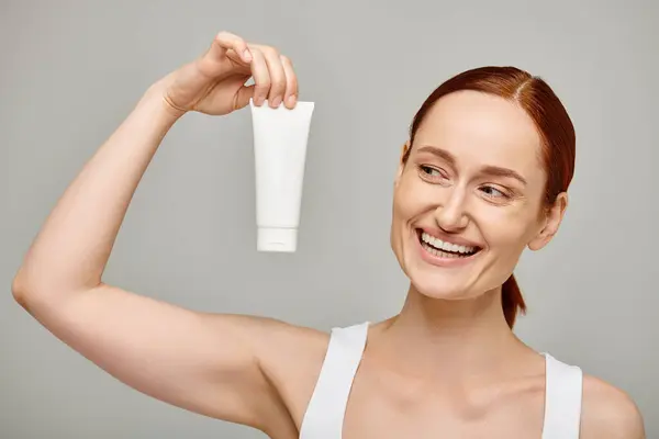 Redhead and cheerful woman in 30s showing tube with body lotion on grey background, skincare — Stock Photo