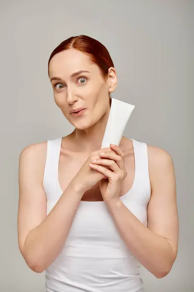 Stupito giovane donna con i capelli rossi che mostra tubo con lozione corpo su sfondo grigio, cura della pelle — Foto stock