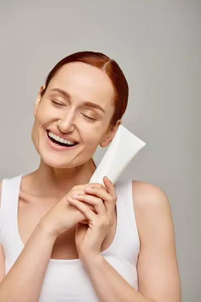 Redhead young and happy woman in 30s holding tube with lotion on grey background, closed eyes — Stock Photo