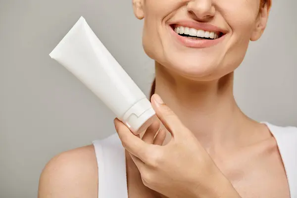 Cropped view of cheerful woman holding tube with body lotion on grey background, skincare — Stock Photo