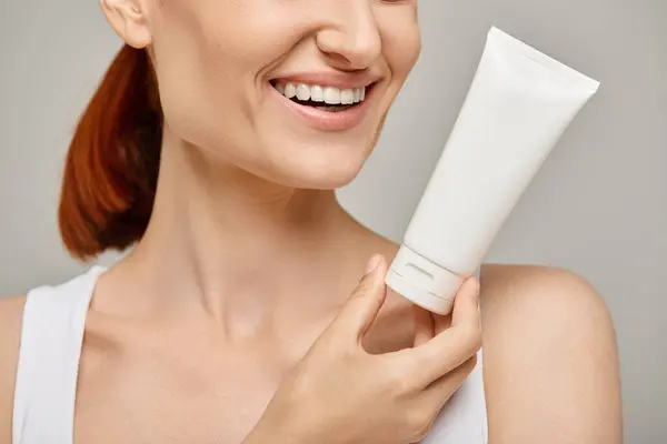 Cropped view of cheerful woman with red hair holding tube with body lotion on grey background — Stock Photo