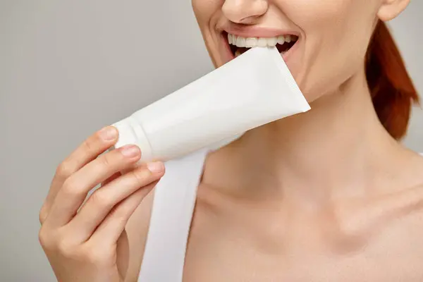 Cropped view of cheerful woman with red hair biting tube with body lotion on grey background — Stock Photo